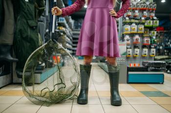 Fisherwoman in rubber boots holds net in fishing shop, hooks and baubles on background. Equipment and tools for fish catching and hunting, accessory choice on showcase in store, bait assortment