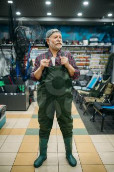 Fisherman tries on rubber jumpsuit in fishing shop, hooks and baubles on background. Equipment and tools for fish catching and hunting, accessory choice on showcase in store