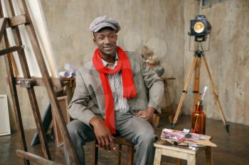 Fashionable male painter in hat and red scarf poses at easel in art studio. Artist draws at his workplace, creative master works in workshop