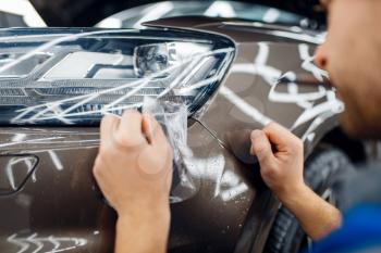 Male worker installs transparent protection film on car headlight. Installation of coating that protects the paint of automobile from scratches. New vehicle in garage, autotuning