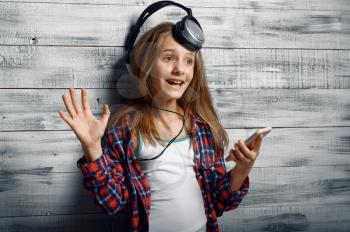 Cheerful little girl in headphones listen to music in studio. Children and gadget, kid isolated on wooden background, child emotion, schoolgirl photo session