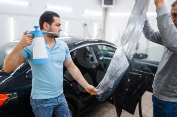 Male worker with spray wetting car tinting, tuning service. Mechanic applying vinyl tint on vehicle window in garage, tinted automobile glass