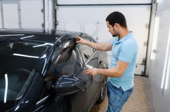 Male worker tries on wetted car tinting, tuning service. Mechanic applying vinyl tint on vehicle window in garage, tinted automobile glass