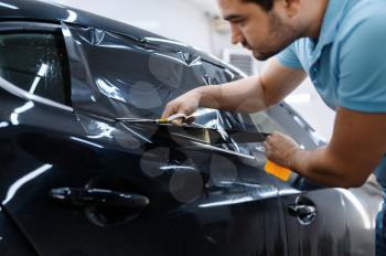 Male worker holds sheet of film, car tinting installation, tuning service. Mechanic applying vinyl tint on vehicle window in garage, tinted automobile glass