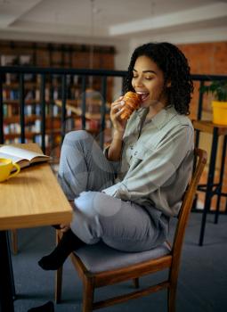Cute female student eats croissants in cafe. Woman learning a subject in coffeehouse, education and food. Girl studying in campus cafeteria