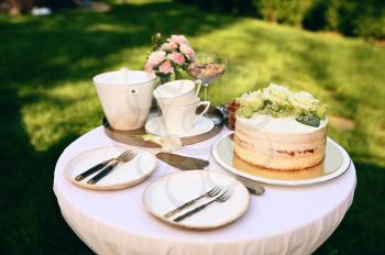 Table setting, ceramic teapot, teacups, cake and flowers closeup, nobody. Luxury silverware on white tablecloth, tableware outdoors