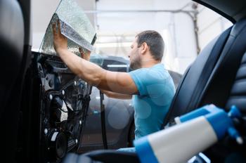 Male specialist installs wetted car tinting, tuning service. Mechanic applying vinyl tint on vehicle window in garage, tinted automobile glass