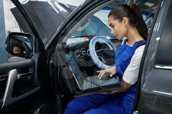 Female mechanic with laptop does the engine diagnostics, car service, professional inspection. Vehicle repairing garage, woman in uniform, automobile station interior on background