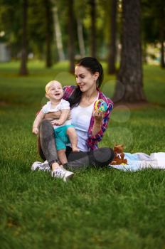 Mother and her little baby poses on grass in summer park. Mom with male kid on lawn, picnic with child on plaid in the forest, family happiness