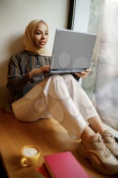 Arab girl in hijab sitting on windowsill and using laptop, university cafe interior on background. Muslim woman with books sitting in library. Religion and education