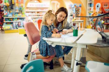 Mother and little baby draws in kid's store. Mom and adorable girl near the showcase in children's shop, happy childhood, family makes a purchase in kid's market