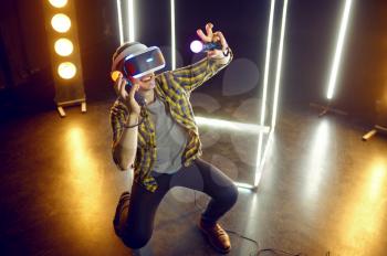 Man gaming in virtual reality headset and gamepad in luminous cube. Dark playing club interior, spotlight on background, VR technology with 3D vision
