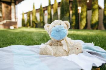 Teddy bear in virus protection mask sitting on a blanket in the garden. Soft toy in respirator on the grass on backyard. Epidemic danger, dangerous infection, health care concept