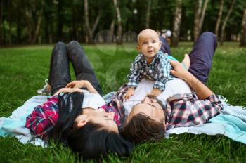 Mother, father and little baby leisures on grass. Mom and dad with male kid on lawn, parents on picnic with child in the forest, family happiness