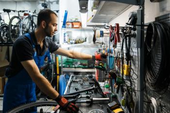 Bicycle repair in workshop, man takes tools from shelf. Mechanic in uniform fix problems with cycle, professional bike repairing service