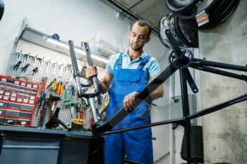 Bicycle assembly in workshop, man installs fork into the frame. Mechanic in uniform fix problems with cycle, professional bike repairing service