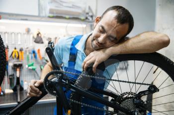 Bicycle repair in workshop, tired repairman sleeps at workplace. Mechanic in uniform fix problems with cycle, professional bike repairing service