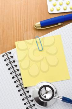 medical stethoscope with pills and notebook on wood