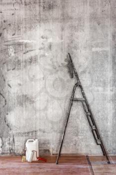 Home interior room repair concept - old gray concrete wall and dirty brown floor in repairing room with old dirty black stepladder near the wall, white jerrican, brush and paint roller,  closeup view 