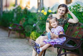 Happy loving family. Mother and little daughter sit on a park bench on a background of bright green foliage.