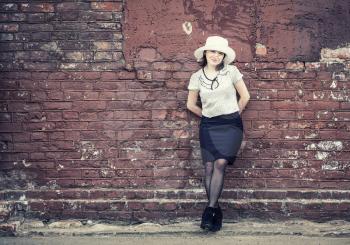 Photo of pretty young woman in white hat, blouse and black skirt, posing against brick wall background. Woman is leaning against the wall. Toned photo with copy space. Vintage style photo.