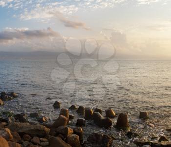 Morning seascape. Calm sea, cloudy sky and mountains on the horizon. Stones and sea walls in the foreground.