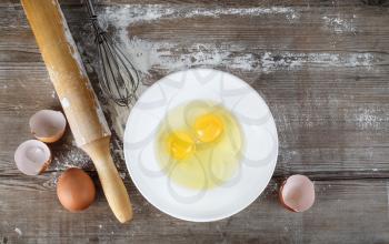 Cooking background with eggs, raw eggs in a dish, eggshells, flour, rolling pin and whisk on a table. Wooden background. Top view.