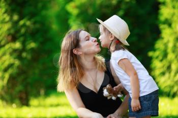 Mom kisses her little daughter. Mom and child girl. Happy family. Bright green foliage in the background. Sunny summer day. Shallow depth of field. Selective focus.