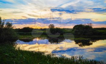 Sunset over the lake, overgrown with sedges. Reflection of the sky and bushes in the water. Evening landscape.