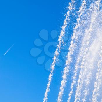 Jets and splashes of water fountain against the blue sky. Condensation track of a jet plane against the sky and a fountain.