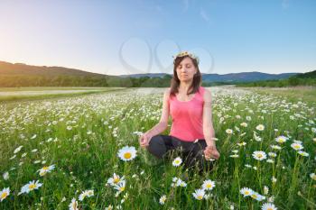 Girl meditates in lotus pose on green camomile meadow. Practicing of yoga at summer. Portrait of young peaceful woman. Training and meditation outdoor. Healthy lifestyle.