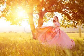 Young woman standing on a wheat field with sunrise on the background. Beauty Romantic Girl Outdoors. The dress fluttering in the wind at sunset. flying fabric.
