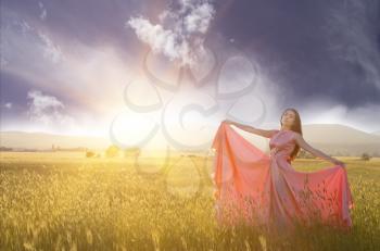 Young woman standing on a wheat field with sunrise on the background. Beauty Romantic Girl Outdoors. The dress fluttering in the wind at sunset. flying fabric.
