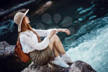 A beautiful young woman with red hair, in a hat and with a backpack near a noisy river in the forest. The idea and concept of freedom, rest, vacations, travel