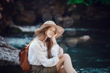 A beautiful young woman with red hair, in a hat and with a backpack near a noisy river in the forest. The idea and concept of freedom, rest, vacations, travel