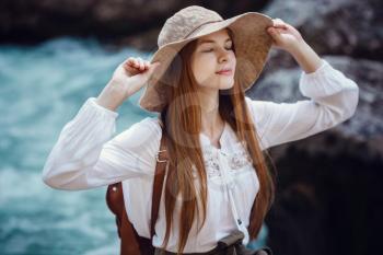 A beautiful young woman with red hair, in a hat and with a backpack near a noisy river in the forest. The idea and concept of freedom, rest, vacations, travel