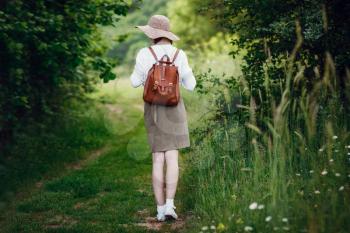 Lovely young woman with red hair, in a hat and with a backpack on the edge of the forest. Beautiful red-haired girl on a walk