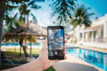 hand holding phone on background of the pool in hotel. photo camera on the screen. close up hand hold smartphone take a picture of summer vacation