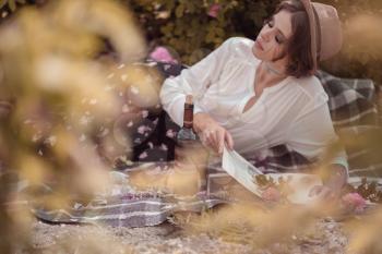 beautiful girl wearing hat with book sitting on grass in rose gaden. A country girl enjoys the sunset, wine and reading books in the fragrant rose garden. The concept of perfume