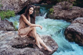 Young woman in the dress is sitting on the stone in the middle of a creek. Fashionable toning.