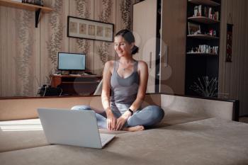 Time for yoga. Attractive young woman exercising and sitting in yoga lotus position while resting at home. View of a woman conducting virtual fitness class at home on a video conference.