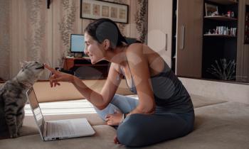 Young attractive smiling woman practicing yoga, sitting in Half Lotus exercise, wearing sportswear, meditation session, indoor full length, home interior, cat near.