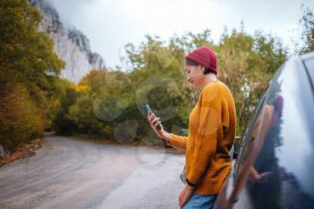 Woman beside a car and looking at smart phone surrounded autumn forest in mountains. Portrait of romantic hipster female, Warm autumn weather, calm scene. Wanderlust photo series.