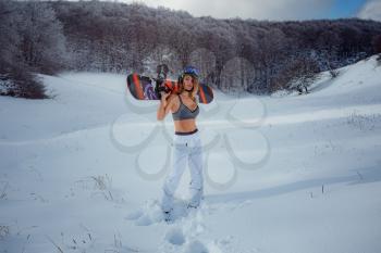 Female snowboarder hold snowboard and going to snowboarding. winter sport activity, forest snow outdoors lifestyle. Girl wearing a short top and ski white pants.