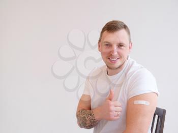 Portrait of young vaccinated man with plaster on his shoulder. Guy gesturing thumb up after coronavirus vaccination, approving of covid-19 immunization.