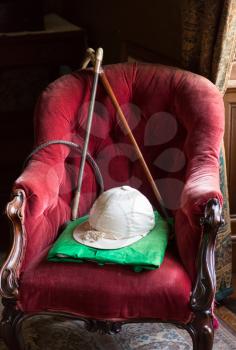 Old horse riding silks and hat with whip and stick on red velvet chair in window light