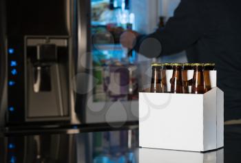 Six pack of brown beer bottles in plain white cardboard carrier with copy space on stainless steel kitchen or bar counter. Open fridge or refrigerator out of focus in rear.