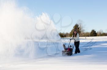 Young lady using a snowblower on rural drive on windy day with a cloud or blizzard of snow blowing in the air