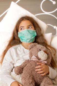 A little girl with her favorite toy bear is lying sick in bed with a gauze bandage over her face.