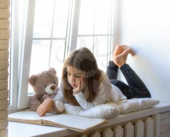 Little Girl lies on the windowsill next to her favorite toy bear cub and reads an old big book